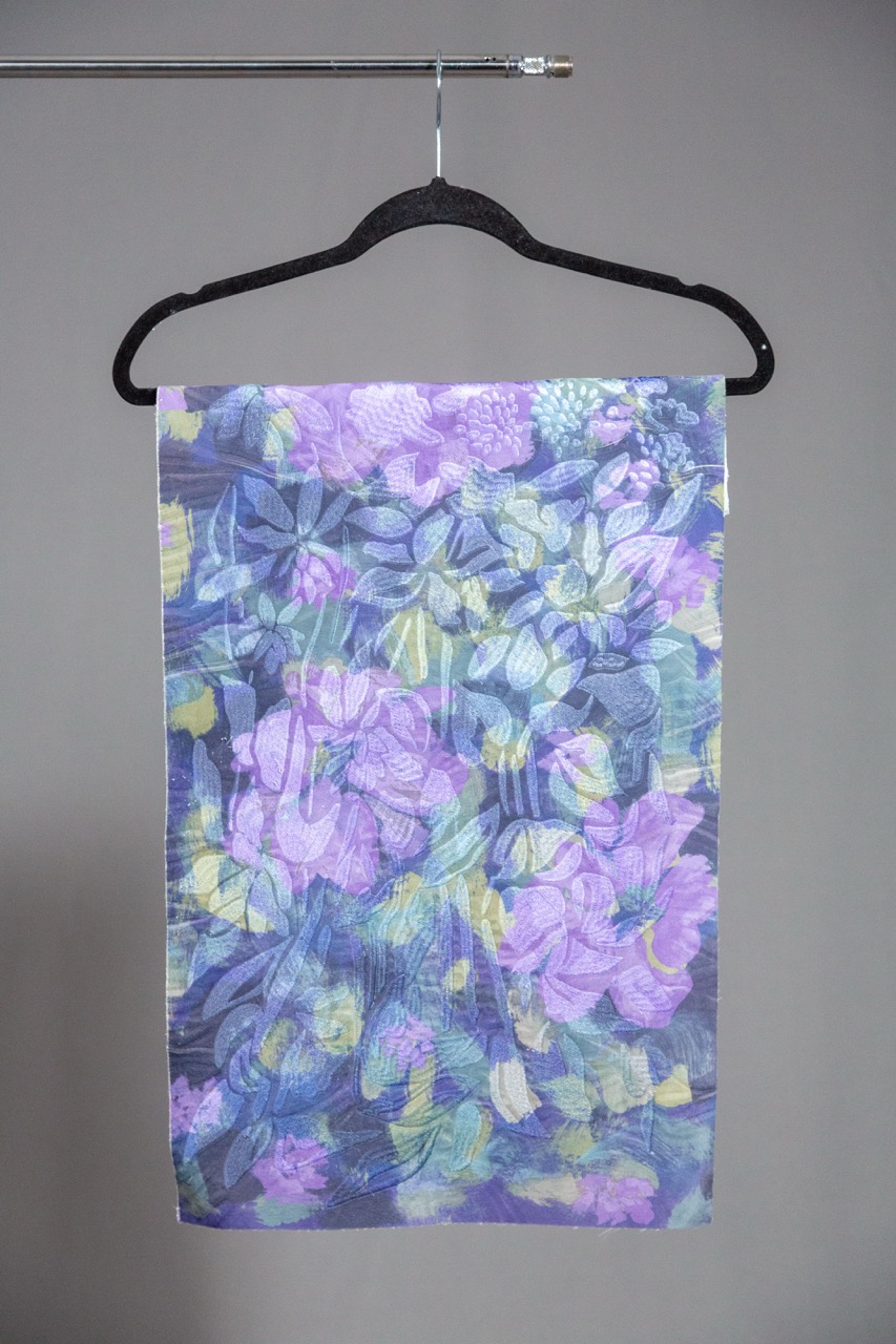 A multicoloured scarf featuring a floral pattern in shades of purple, blue, and yellow. The scarf is hanging from a black clothes hanger against a grey background.