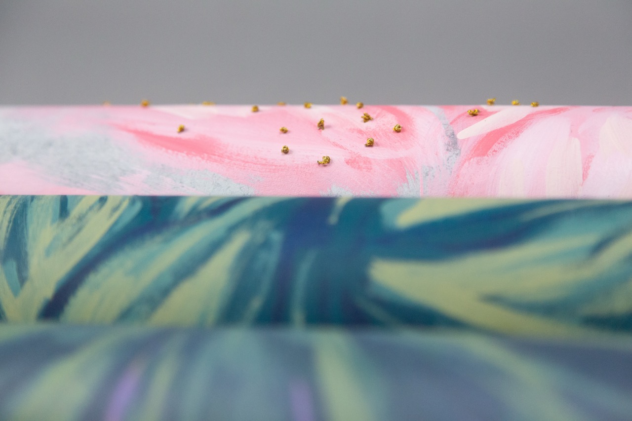 Three rolls of decorative paper are stacked with varying designs. The top roll features a pink and white marbled pattern with small gold embellishments, while the middle roll has a blue and green abstract design, and the bottom roll shows a solid grey background.