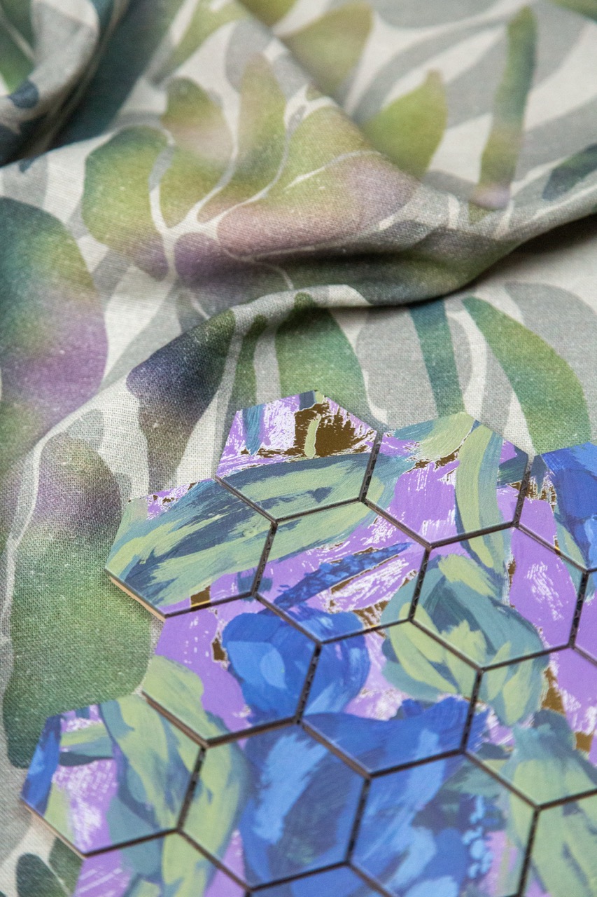 A close-up view of a textured fabric featuring a floral design in green and purple tones, with an overlay of hexagonal tiles in shades of blue and purple, arranged against a soft, patterned background.