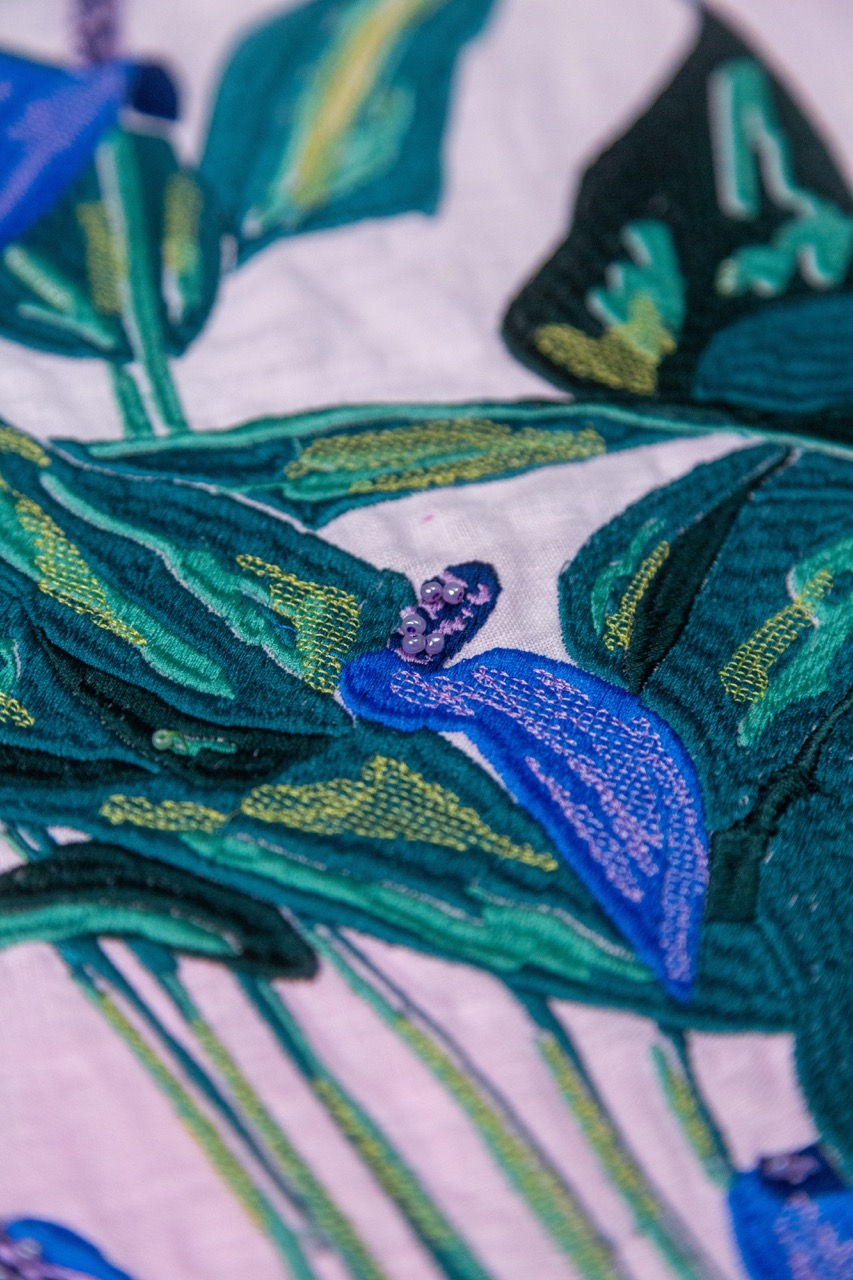 A close-up view of embroidered floral patterns featuring blue flowers and green leaves on a light background. The intricate stitching highlights the different textures and shades of the design.