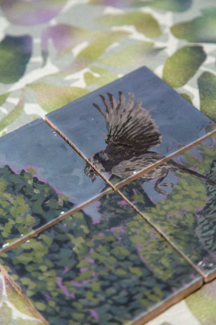 Four ceramic tiles arranged together, featuring a painted bird in flight above a green landscape with purple flowers. The background has a soft, blurred leaf pattern, enhancing the focus on the tiles.