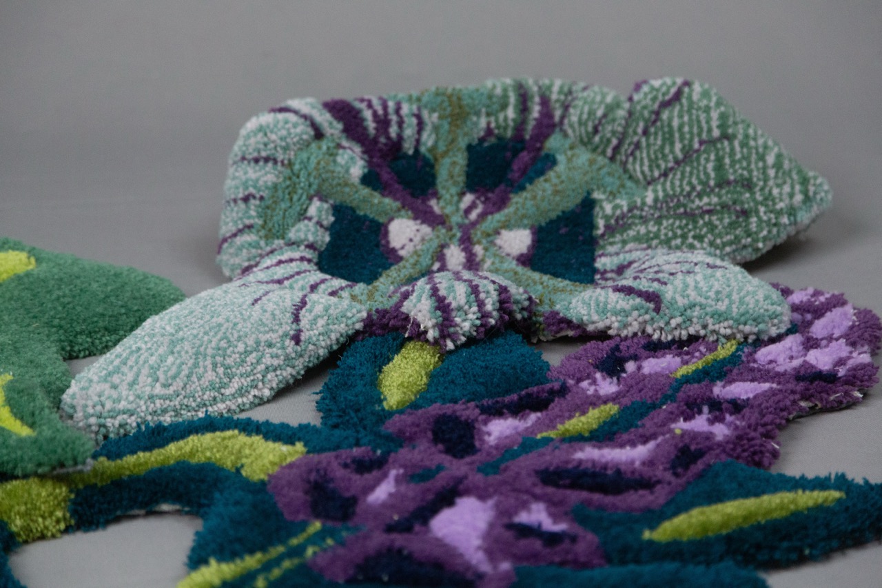 A close-up of a textured carpet featuring floral designs in shades of green, purple, and blue. The pattern includes overlapping petals and leaves, with a mix of yarn-like materials creating a dimensional appearance.