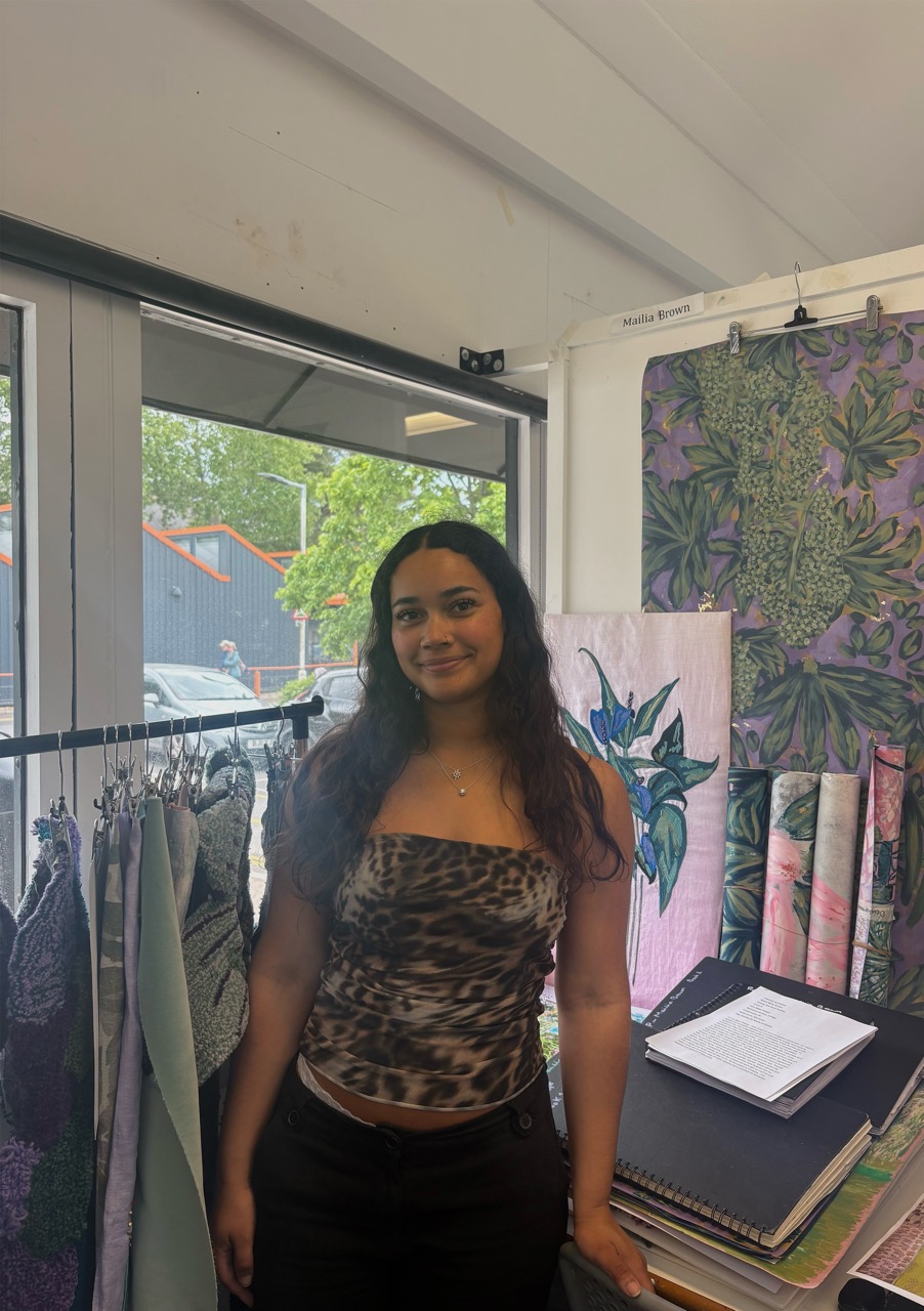 A woman wearing a leopard-print top stands next to draped fabric samples in a bright space. Behind her, large windows showcase greenery and a building. Artworks with floral designs are visible nearby on a table.