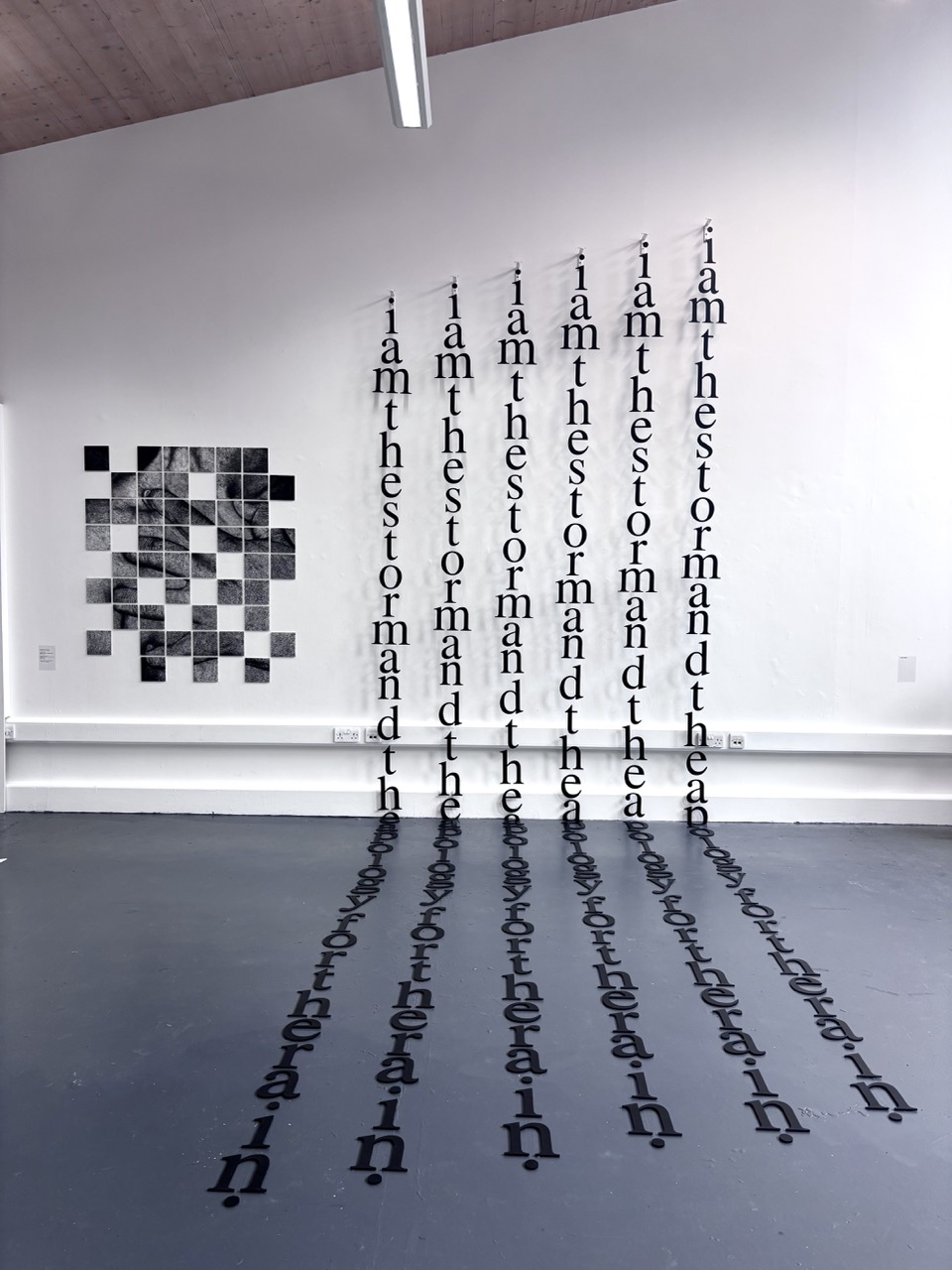 A contemporary art installation featuring text reading "i am the storm and the" arranged vertically on a white wall. Below, the same text casts shadows on the grey floor, while a patterned square design is displayed on the left side of the wall.