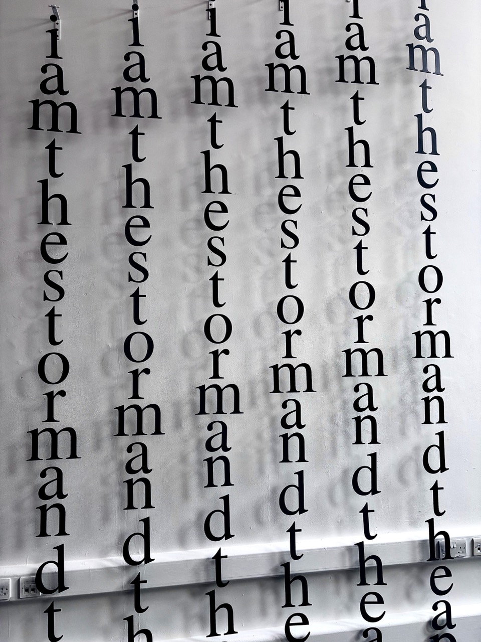 Black letters arranged vertically on a white wall, spelling out the phrase "I am the storm and the" repeatedly in varying font sizes. The letters are hung in a staggered pattern, creating a rhythmic visual effect.
