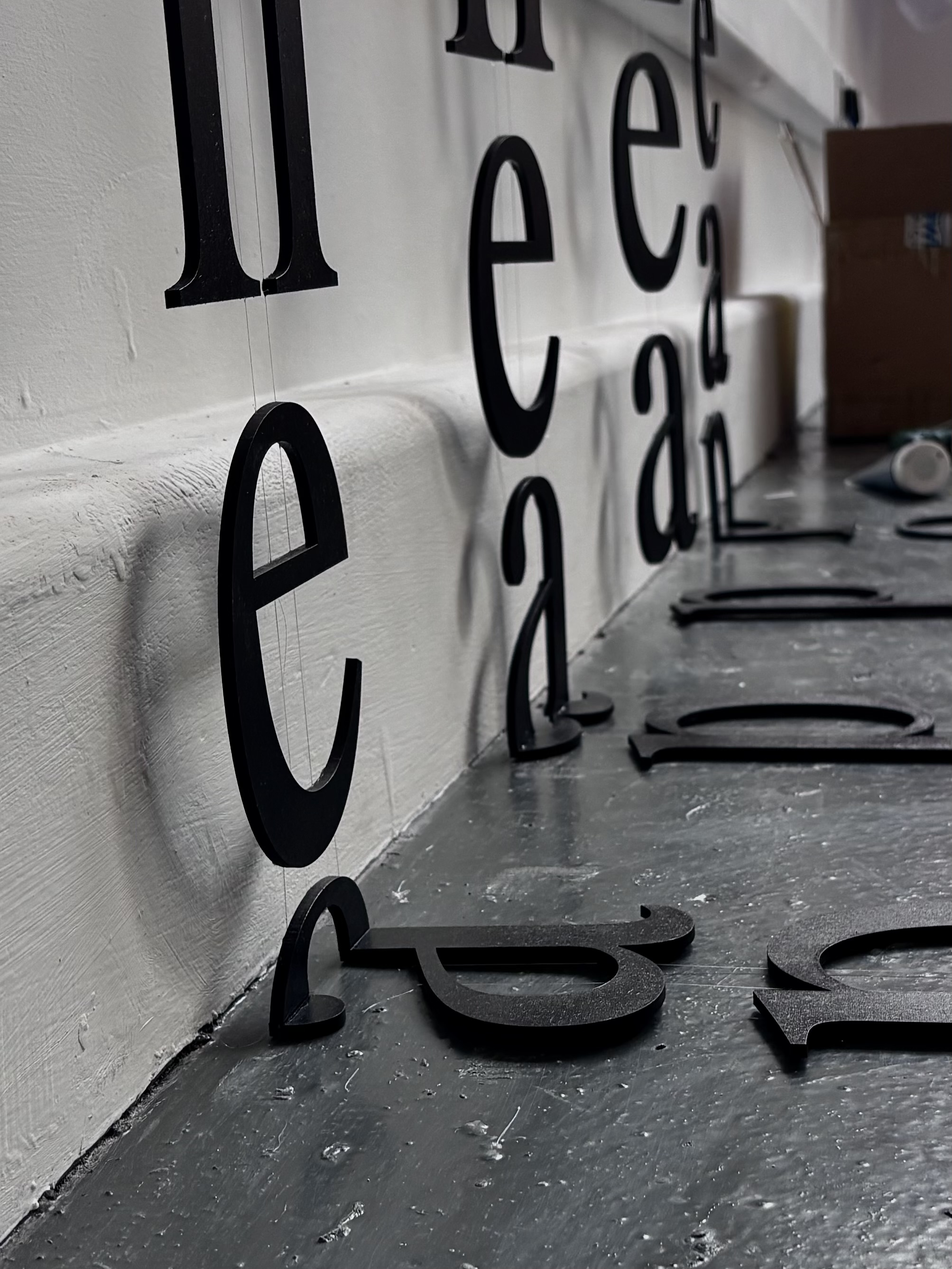 A collection of black wooden letters arranged on a grey floor, with some letters leaning against a white wall. The lower half of letters such as 'e' and 'a' are clearly visible, highlighting their shapes and design.