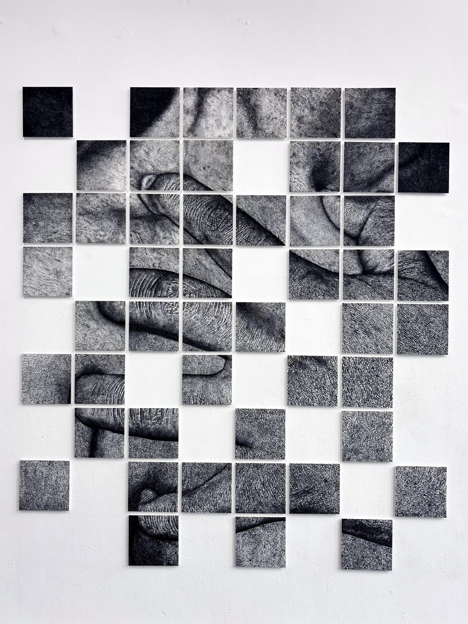 An artwork composed of multiple square tiles arranged in a grid pattern, displaying black and white images of hands and skin texture. The tiles vary in tones, creating a fragmented yet cohesive visual representation.