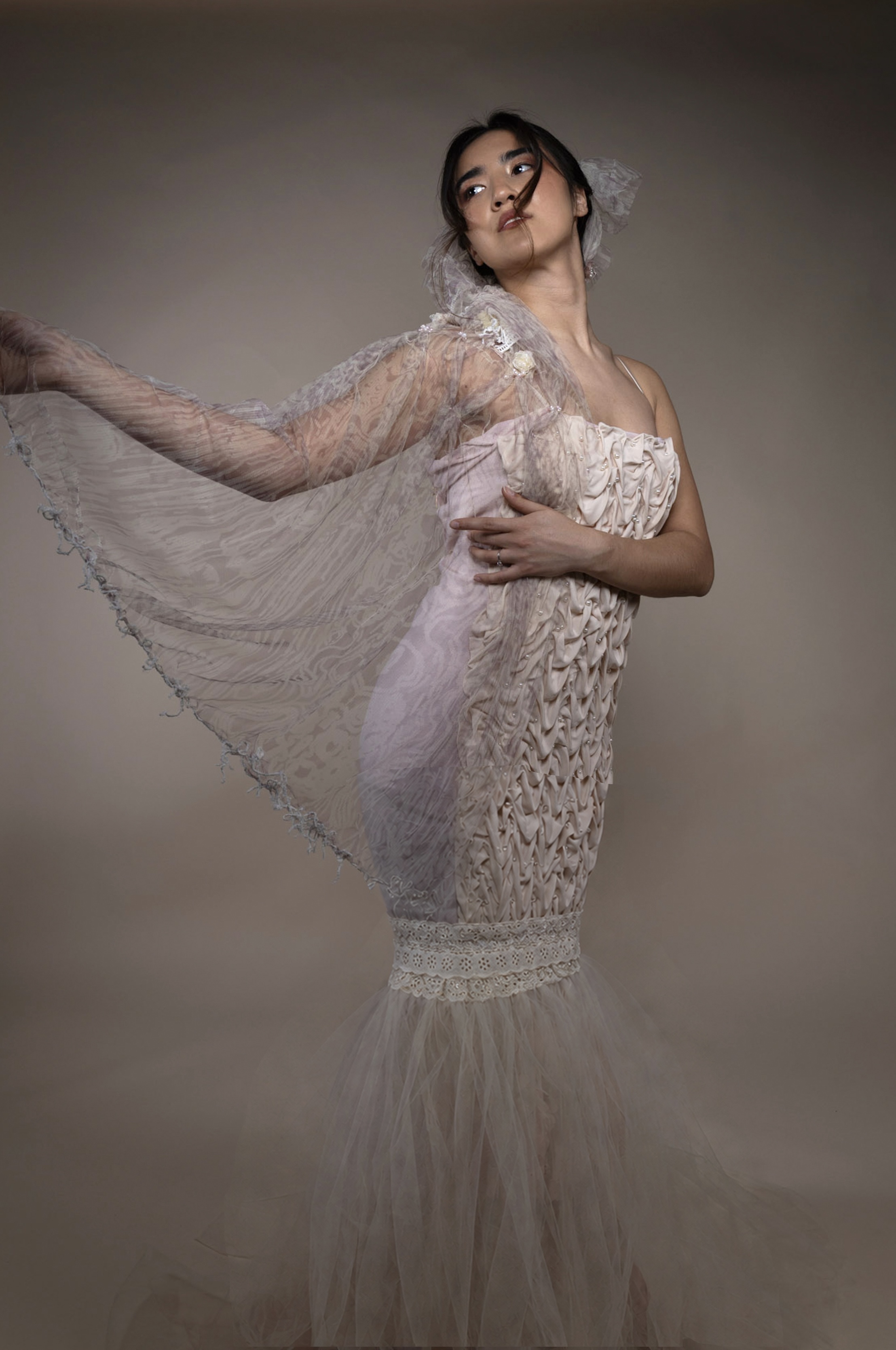 A woman wearing a fitted, strapless wedding dress made of soft fabric and lace, with a flowing veil draped over her shoulder. The background is a muted tone, enhancing the delicate details of the dress and the woman's pose.