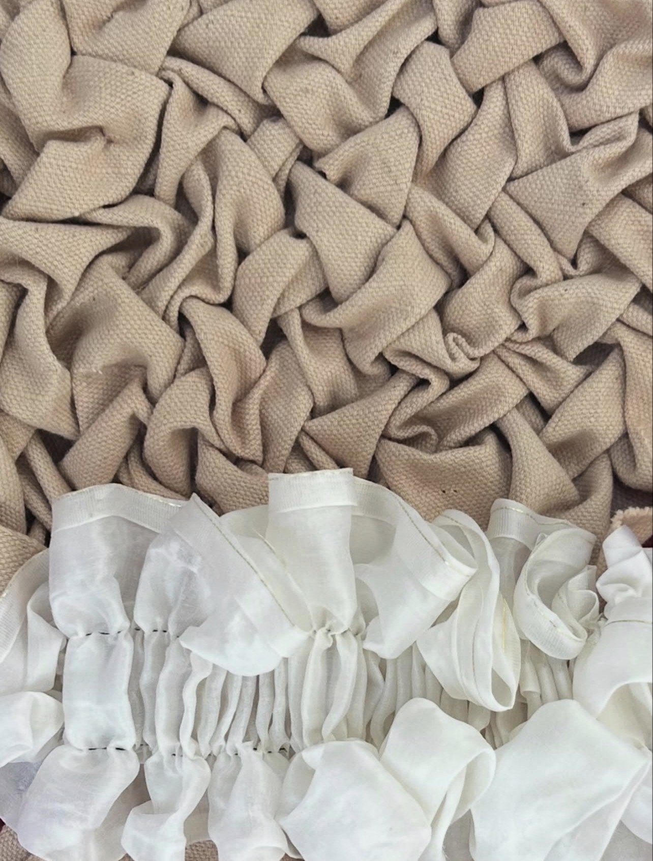 A textured surface featuring crumpled beige fabric creating a wavy pattern, with a section of white, wrinkled fabric at the bottom displaying gathered ruffles. The two fabrics contrast in colour and texture.