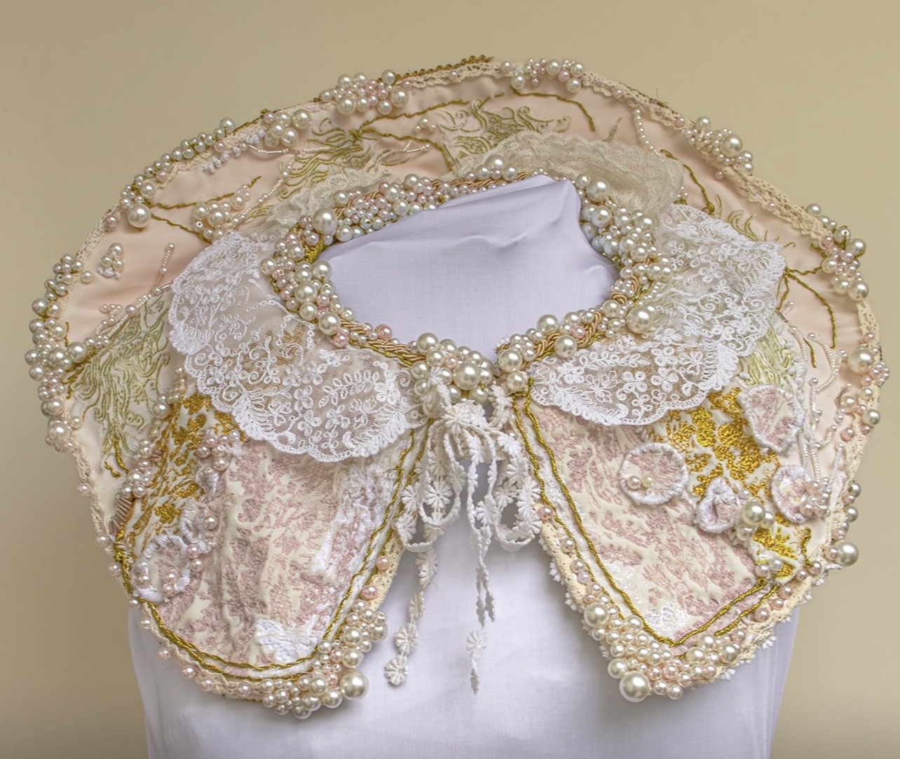 An ornate collar displayed on a mannequin, featuring white lace trim, gold embroidery, and intricate beadwork. The collar has a pastel pink fabric base adorned with floral patterns and is embellished with pearls and sequins.