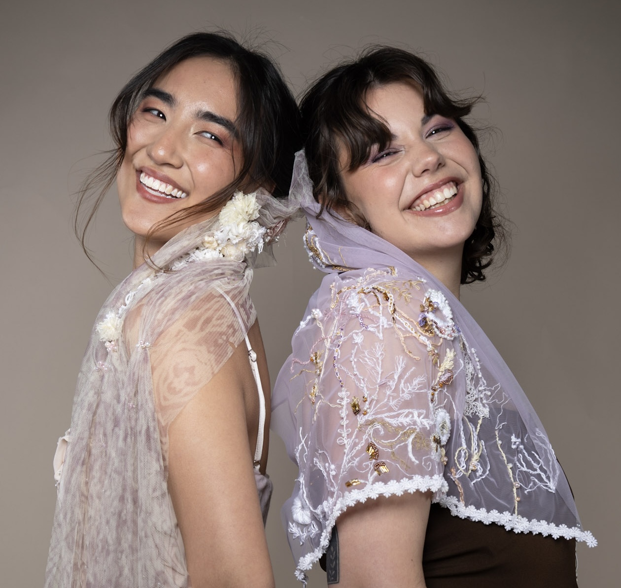 Two women stand back to back, smiling. The woman on the left wears a sheer, floral-embroidered shawl, while the woman on the right wears a light purple shawl with decorative embellishments. They have dark hair and are against a neutral background.