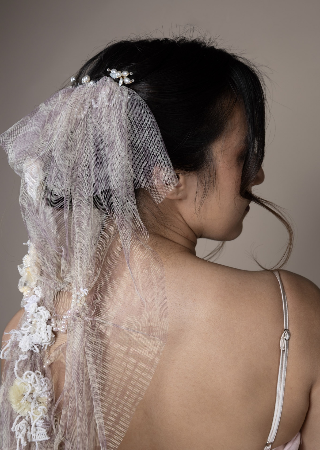 A young woman with long dark hair is viewed from the back, adorned with a delicate veil featuring lace and floral embellishments. She wears a light-coloured spaghetti-strap dress, and the background is a soft beige.