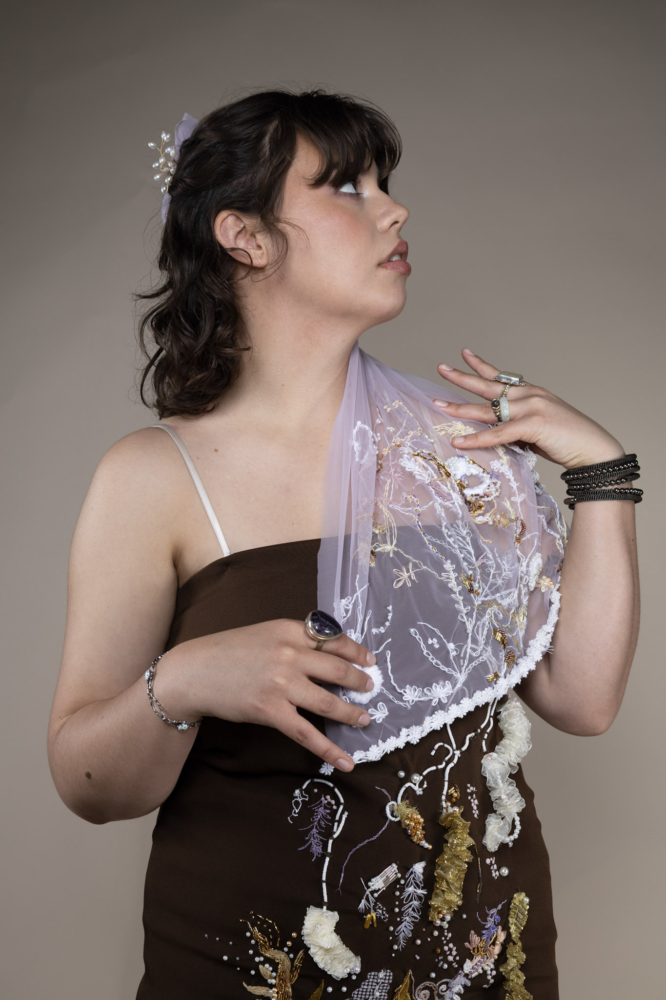 A woman with shoulder-length dark hair and a decorative hairpiece poses wearing a brown garment embellished with floral designs. She holds a sheer, embroidered veil draped over her shoulder, against a neutral background.