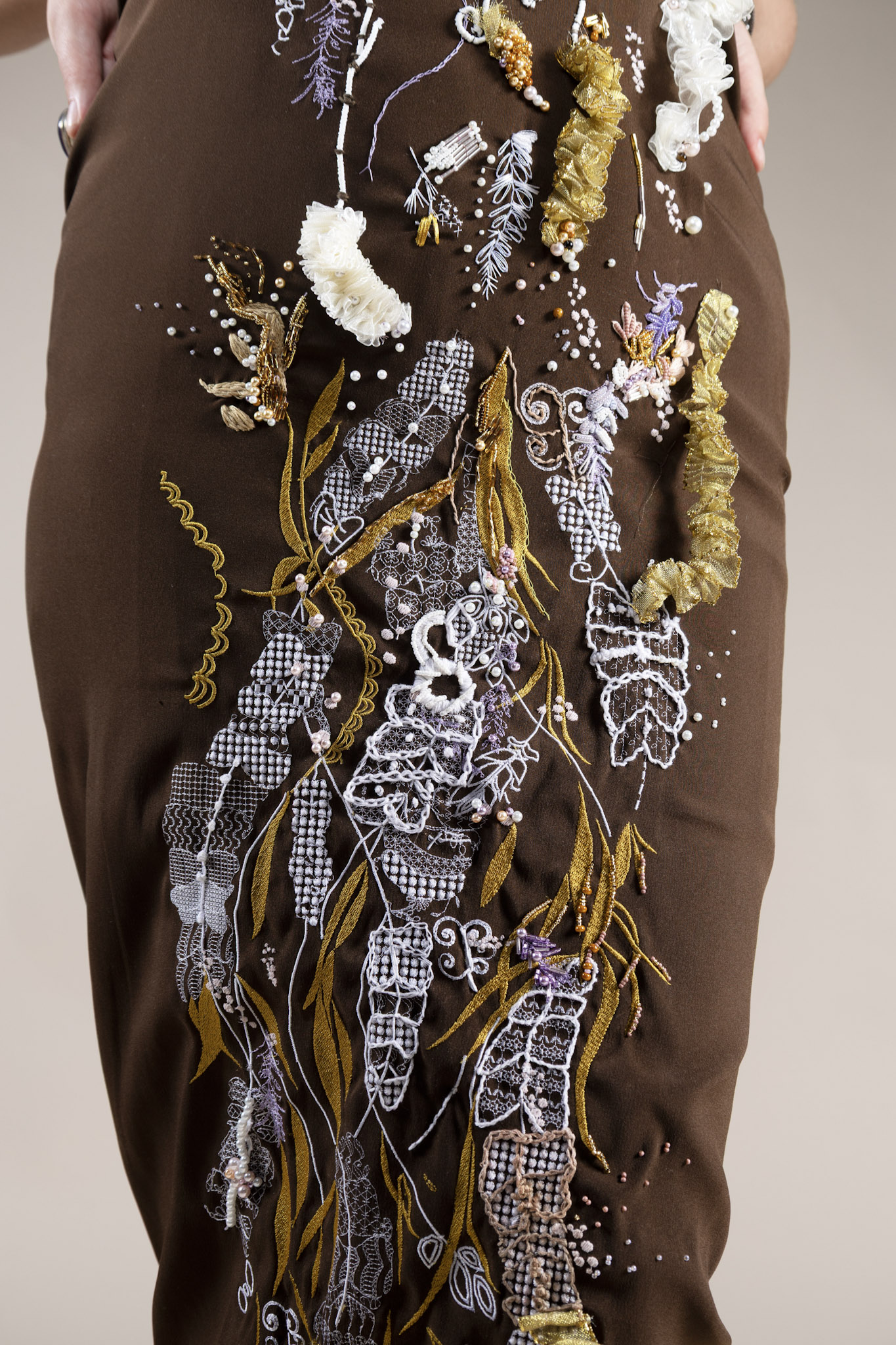 A close-up of a brown garment featuring intricate embroidery. The design includes various floral and abstract patterns in white, purple, and gold threads, showcasing a mix of textures and shapes along the fabric's side.