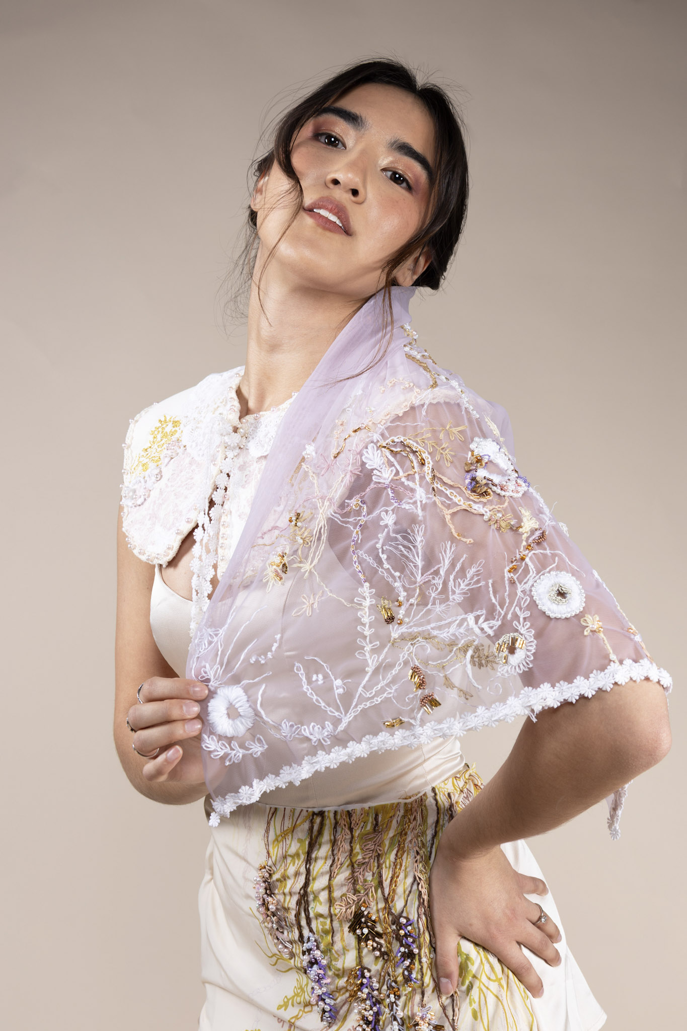 A woman stands in front of a neutral background, wearing a white top with a high collar and a sheer embroidered shawl draped over her shoulders. The shawl features floral patterns and delicate embellishments, while she poses with her right hand on her hip.