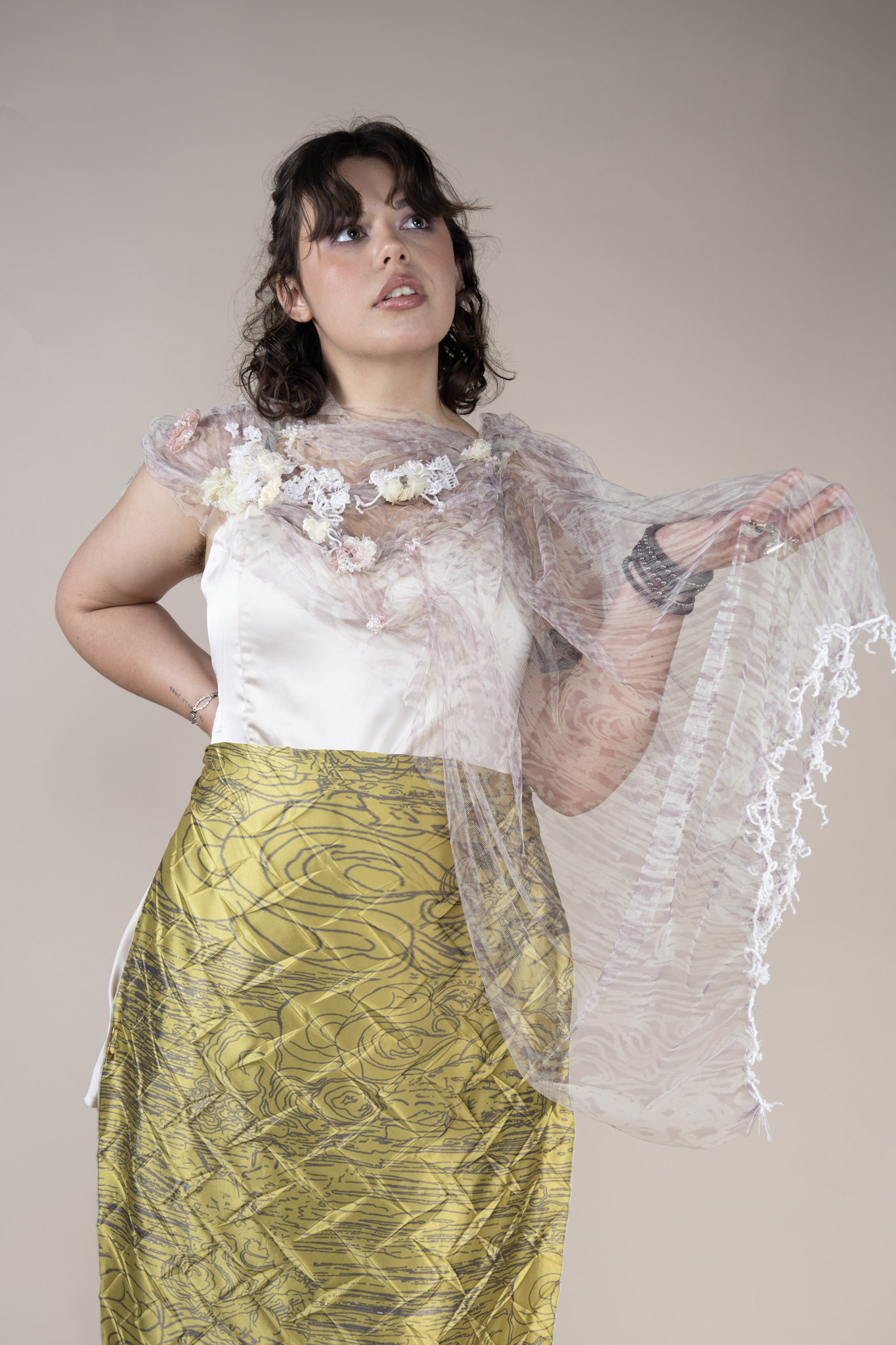 A model poses against a neutral background, wearing a white satin top adorned with floral details and a lightweight, textured shawl. She has a flowing, patterned yellow skirt and holds the shawl elegantly in one hand.
