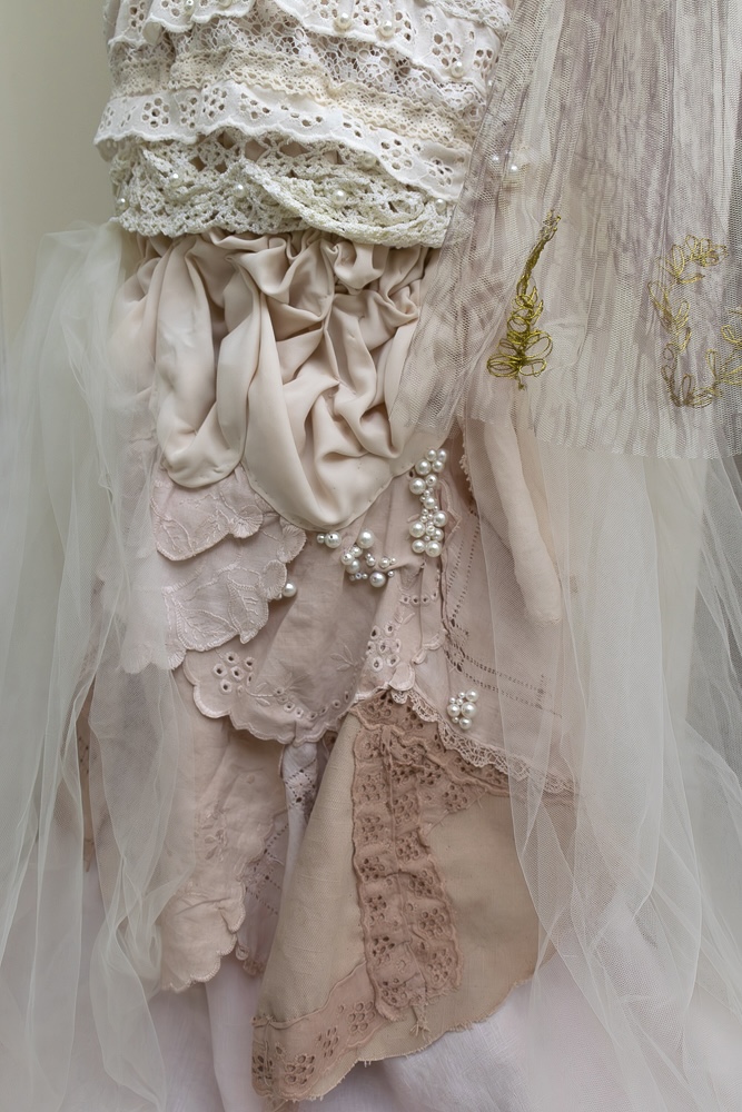 A close-up view of a dress made from various light fabrics, including lace, tulle, and silk in soft pink and cream colours. The material features intricate details, ruffles, and embellishments, such as pearls and delicate stitching.