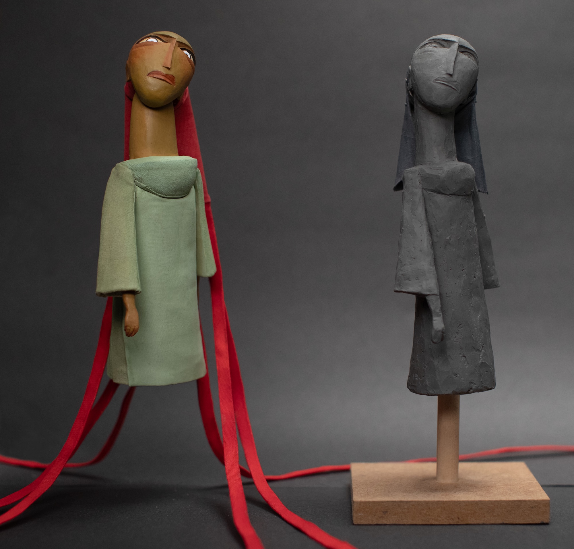 Abstract puppet of a person with long, red hair and serious expression wearing a green garment, sculpture clay version in grey on a wooden stand next to it.