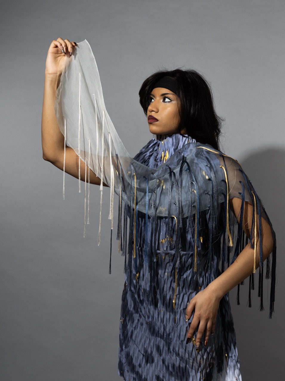 A person wearing a textured grey outfit with gold accents holds a sheer white fabric with long fringes in one hand. The background is a neutral grey, focusing on the individual's pose and the details of the attire.
