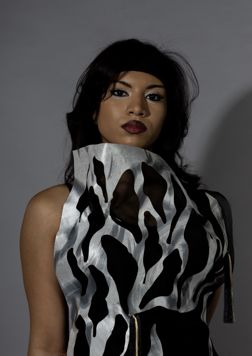 A woman stands against a grey backdrop, wearing a striking dress made of black fabric with silver cutouts in an abstract pattern. Her hair is dark and styled, and she has bold makeup with a focus on her eyes.