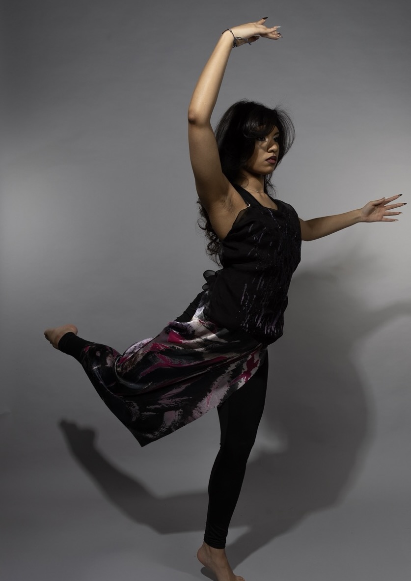 A woman performing a dance pose with her right arm raised and her left leg extended. She is wearing a black sleeveless top and a flowing skirt with a colourful pattern, standing on bare feet against a neutral grey background.