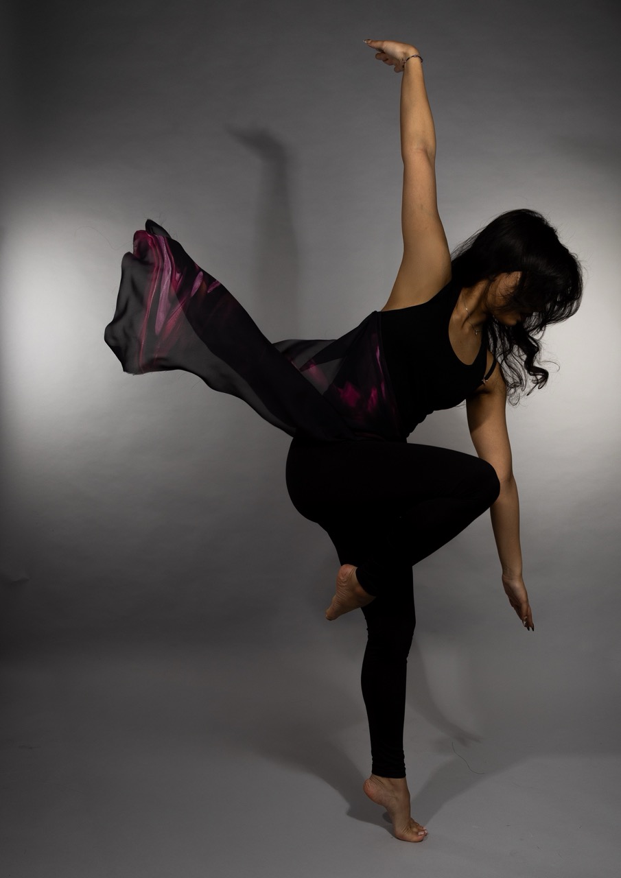 A woman in a black tank top and leggings performs a dance pose, with one arm raised and her right leg extended. She wears a flowing, sheer garment with pink accents that drapes behind her. The background is grey, enhancing the motion of her attire.