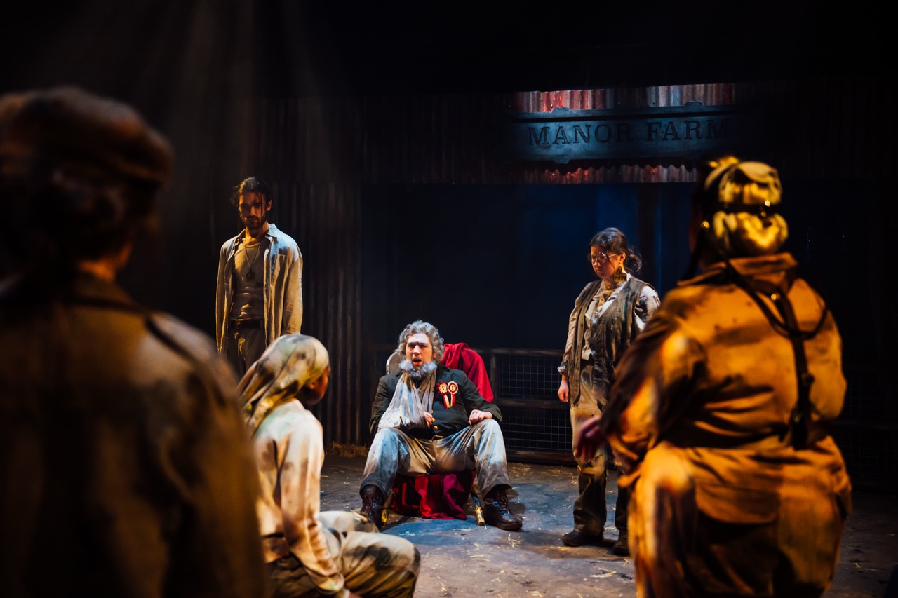 A group of six actors performing on stage in a dimly lit setting. A central figure with white hair, seated and dressed in grey, is surrounded by five standing performers in mismatched, worn clothing, with a backdrop featuring the words "MANOR FARM."
