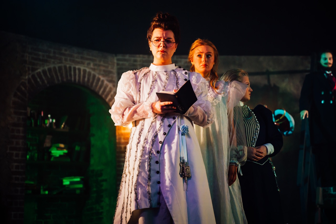 A person dressed in a white historical costume holds a book while standing in front of two women in white dresses. The background features a dimly lit stage with props and a shadowy figure in a dark suit.