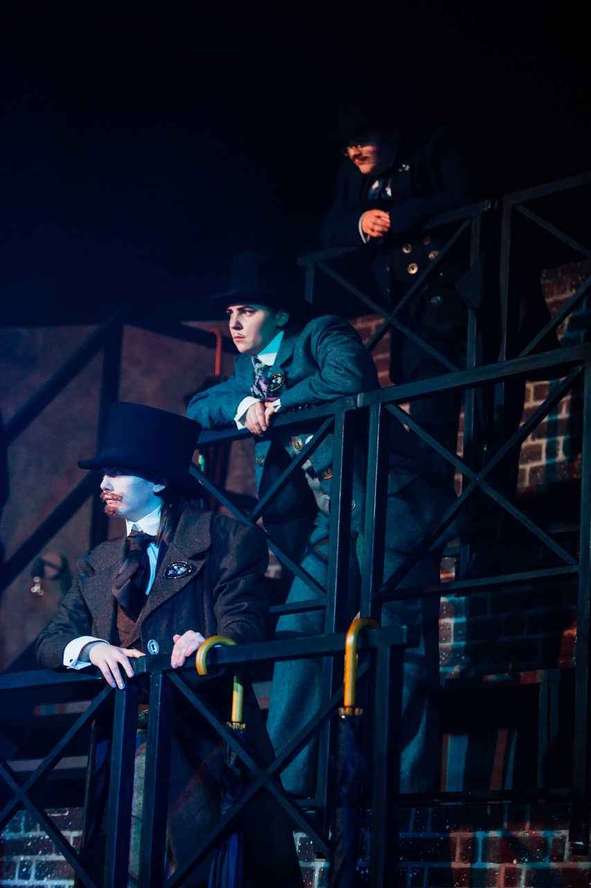 Three men dressed in Victorian-era clothing stand on a metal balcony. One man leans on the railing, while another looks downwards. The background features a brick wall, and the scene is illuminated with dim stage lighting.