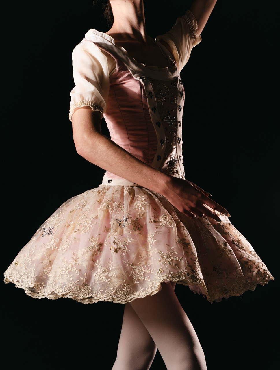 A ballet dancer's silhouette in a pink tutu dress with ornate lace trim and a fitted bodice. The dancer raises one arm gracefully, posed against a dark background that highlights the dress's details.