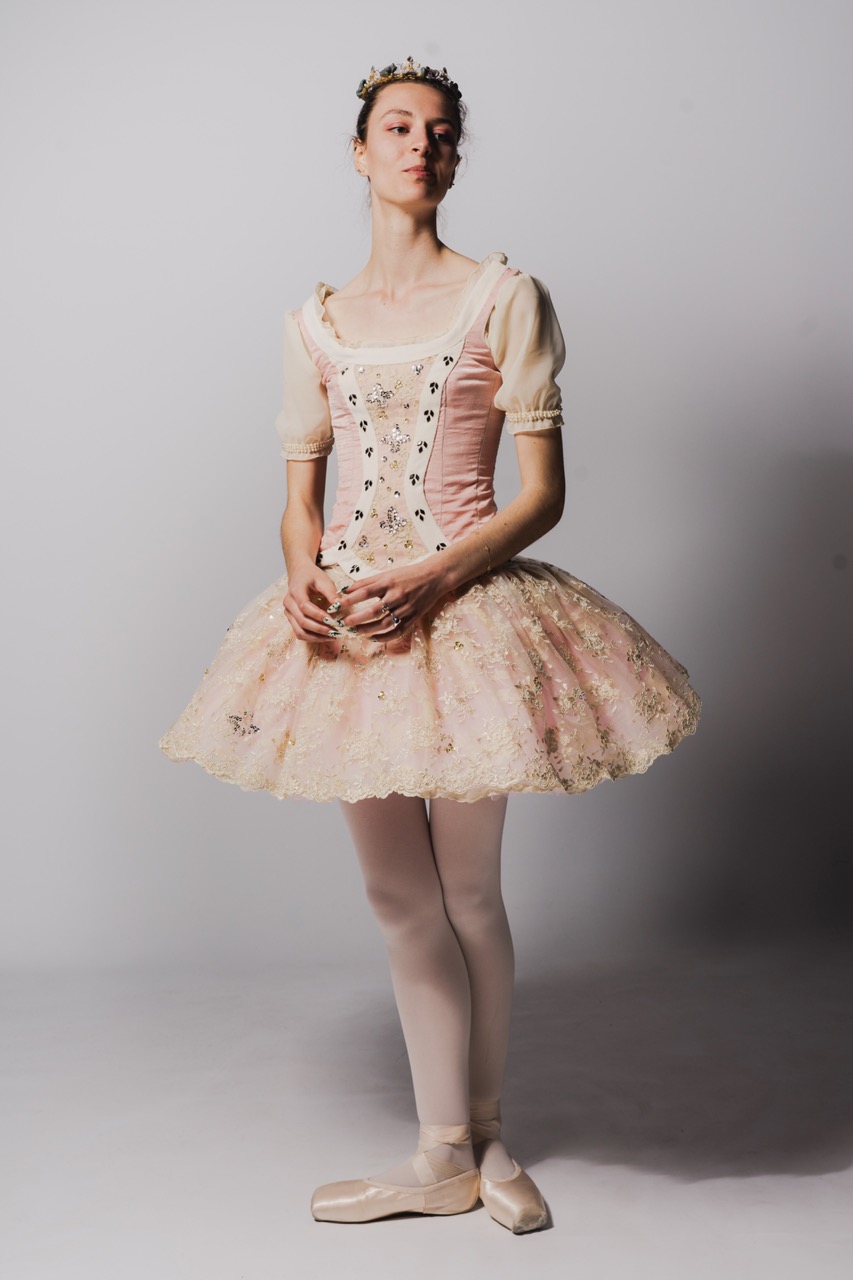 A ballet dancer wearing a pink tutu and a fitted bodice adorned with floral designs. She stands poised with her hands clasped in front of her, wearing pink tights and ballet shoes, against a plain grey background.