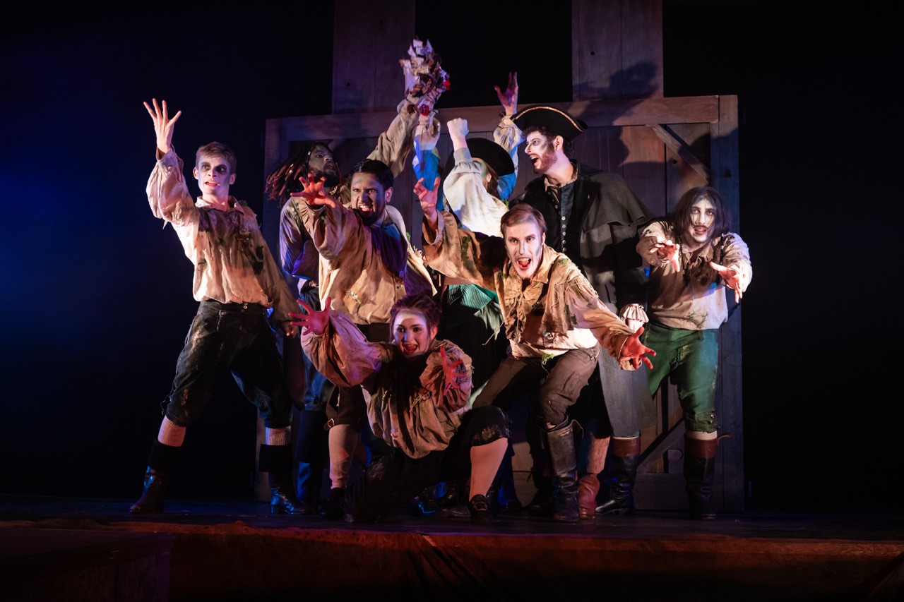 A group of eight actors dressed in tattered costumes, with painted faces, poses dramatically on stage. They stand in front of a wooden backdrop, with dim lighting creating a theatrical effect.