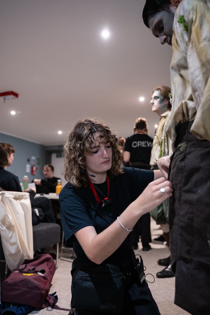 A young woman with curly hair in a black shirt adjusts the trousers of a person dressed in a tattered zombie costume. Behind them, other crew members are visible, along with chairs and a table in a well-lit room.