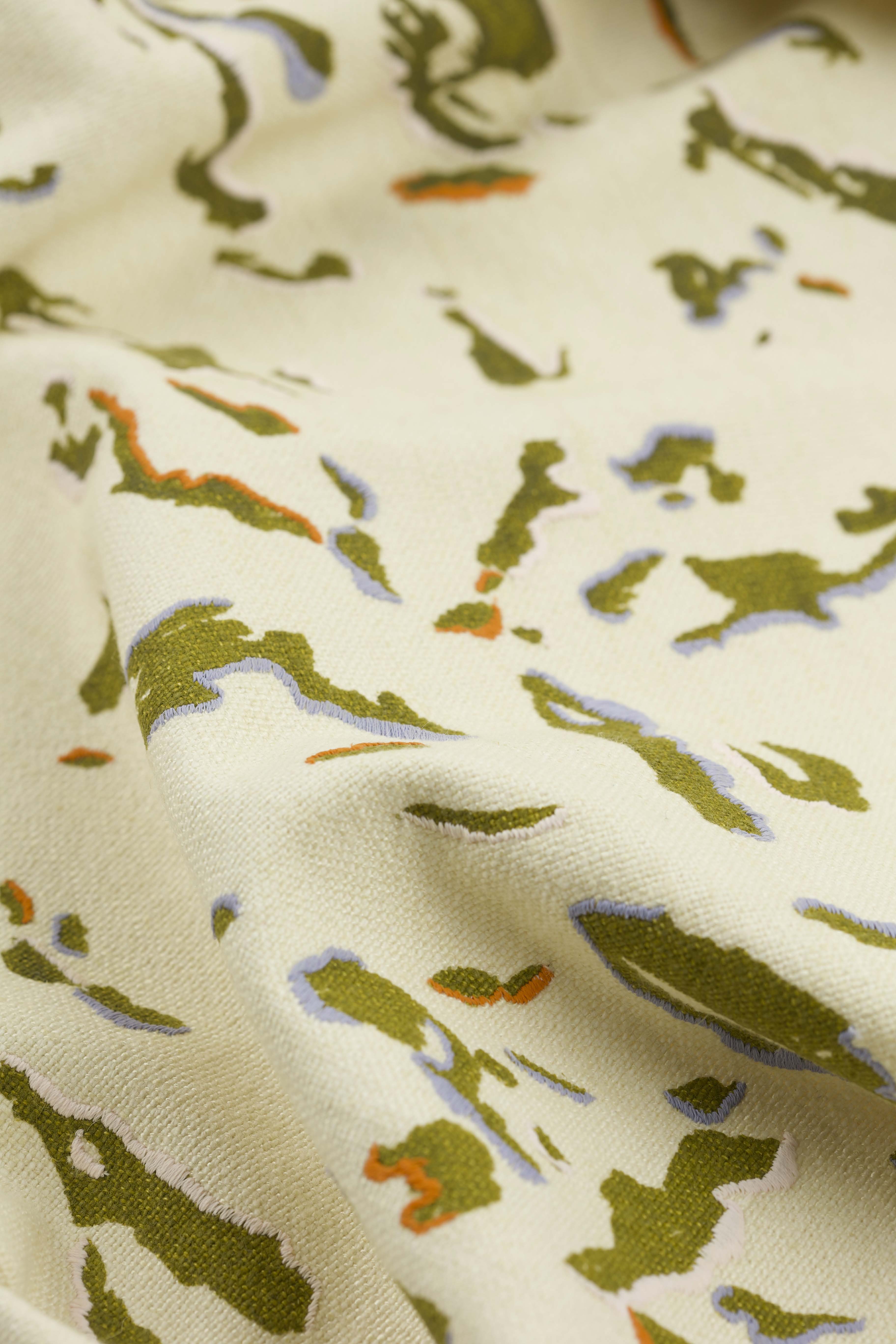 A close-up image of fabric featuring a pattern of abstract shapes in green, orange, and lavender tones on a cream background. The texture of the fabric is soft and appears slightly wrinkled, enhancing the visual depth of the design.