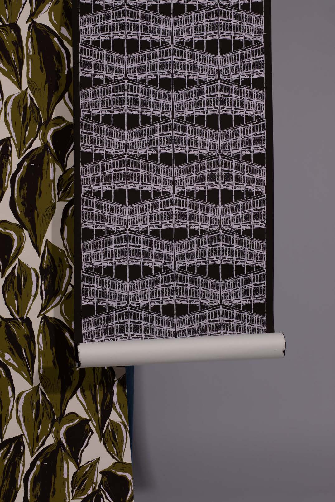 A roll of wallpaper with a geometric white pattern against a black background hanging beside another fabric featuring large green leaves. The wall is a muted grey, providing a neutral backdrop for the designs.