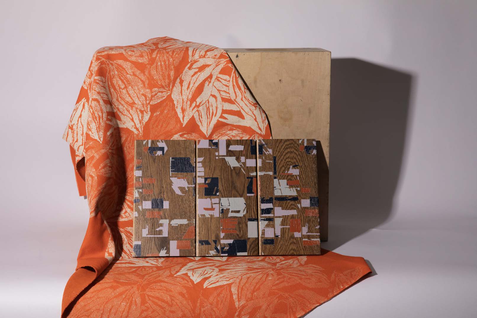 An orange fabric with a leaf pattern drapes over a flat surface, partially covering three wooden panels with an abstract design. Behind them, a plain wooden block rests against a neutral backdrop.