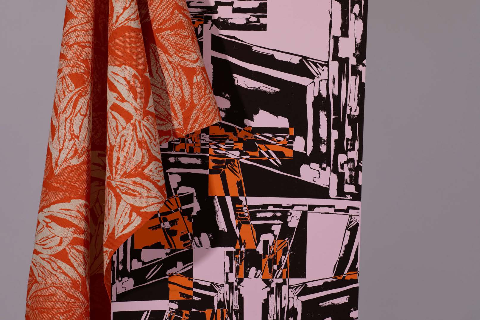 An orange and white patterned fabric drapes over a pink and black abstract patterned textile. The background is a neutral grey, highlighting the contrasting designs and colours of the fabrics.