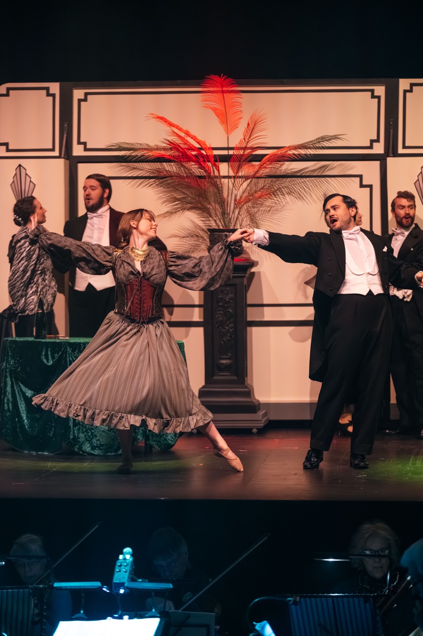 A dancer in a long grey skirt and corset performs on stage, holding hands with a man in a black tuxedo. In the background, other cast members are dressed in formal attire, while a large decorative plant with feathers is positioned centrally.