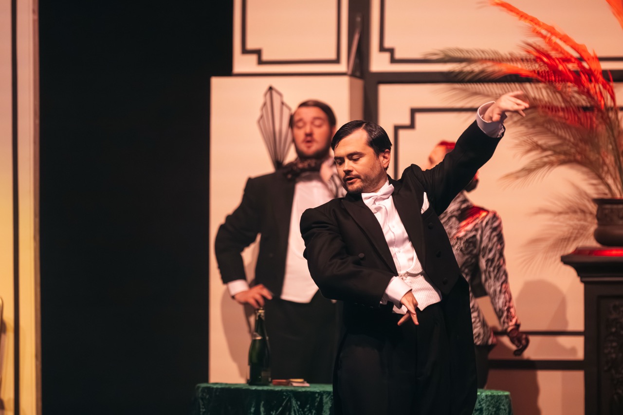 A man in a tuxedo performs on stage, gesturing with his right hand. In the background, another man in a white shirt and bowtie stands beside a decorative display featuring feathers. The setting appears to be a theatrical performance.