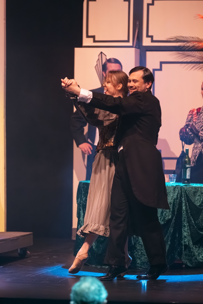 A man in a formal black coat dances with a woman wearing a grey dress on a stage. In the background, several other performers are present, with a table covered in green fabric displaying wine bottles, under theatrical lighting.
