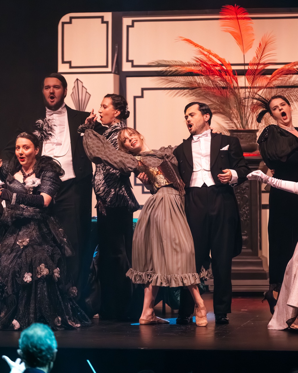 A group of performers in theatrical costumes on stage. Two men in tuxedos are positioned at the front, while women in elegant dresses are arranged behind them, all engaged in a dramatic performance. The backdrop features decorative details and a plant.