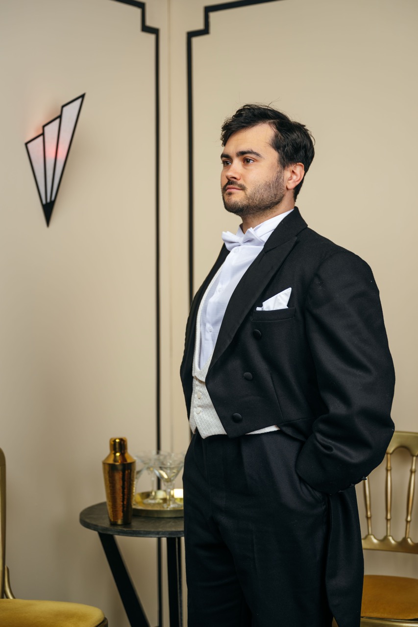 A man dressed in a black tuxedo with a white bow tie and pocket square stands beside a small table with a cocktail shaker and glassware. The background features geometric wall decor and a stylish chair, enhancing the elegant setting.