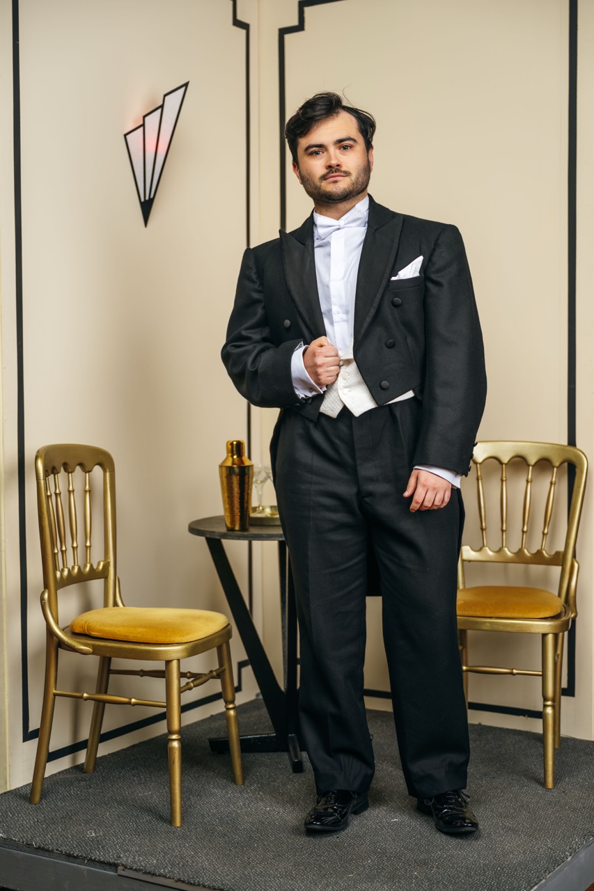 A young man in a formal black tuxedo stands beside a small round table with a gold shaker and glass. Two gold chairs are placed adjacent to the table against a backdrop featuring a geometric design.