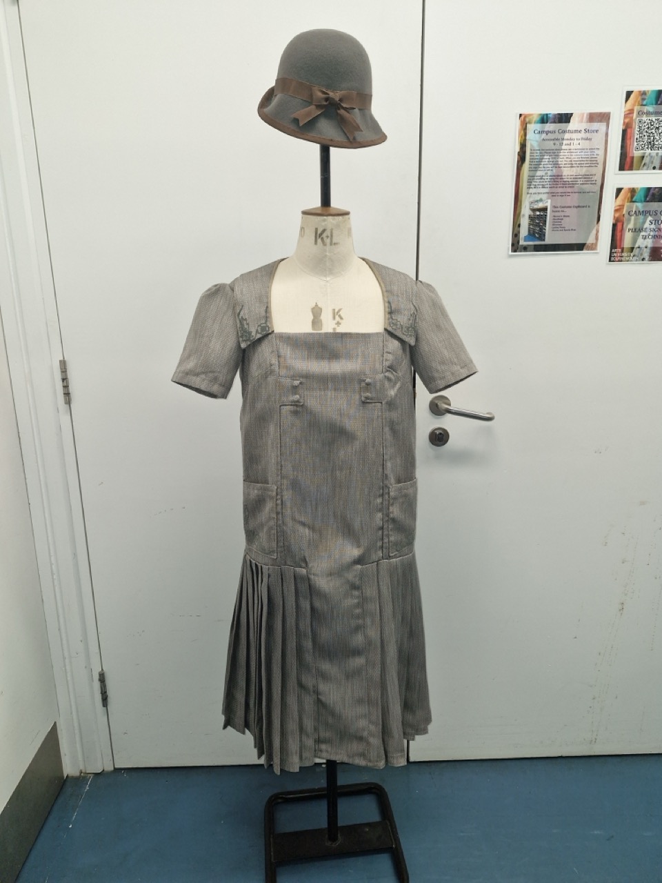 A grey striped dress with short sleeves and a pleated skirt displayed on a dress form. A matching grey hat with a bow sits atop the form. The background features a white wall and several colourful paper displays.