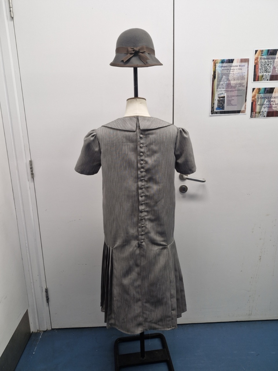 A vintage grey dress displayed on a mannequin, featuring a Peter Pan collar and pleated skirt with buttons running down the back. An accompanying grey cloche hat adorned with a bow rests on top of the mannequin's head.