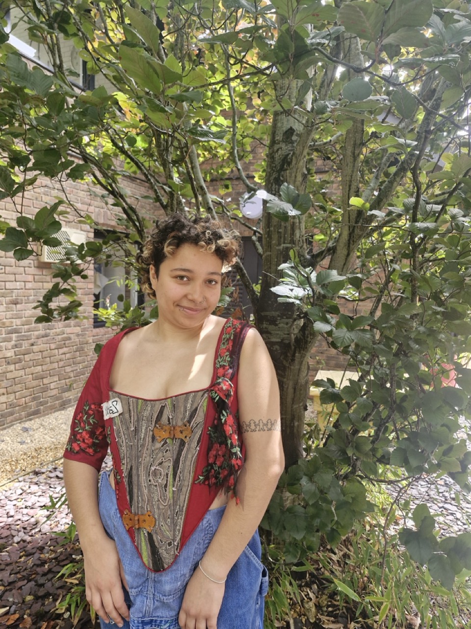 A person with short curly hair stands in front of a tree, dressed in a floral top and denim overalls. The background features brick walls and a pathway lined with pebbles.