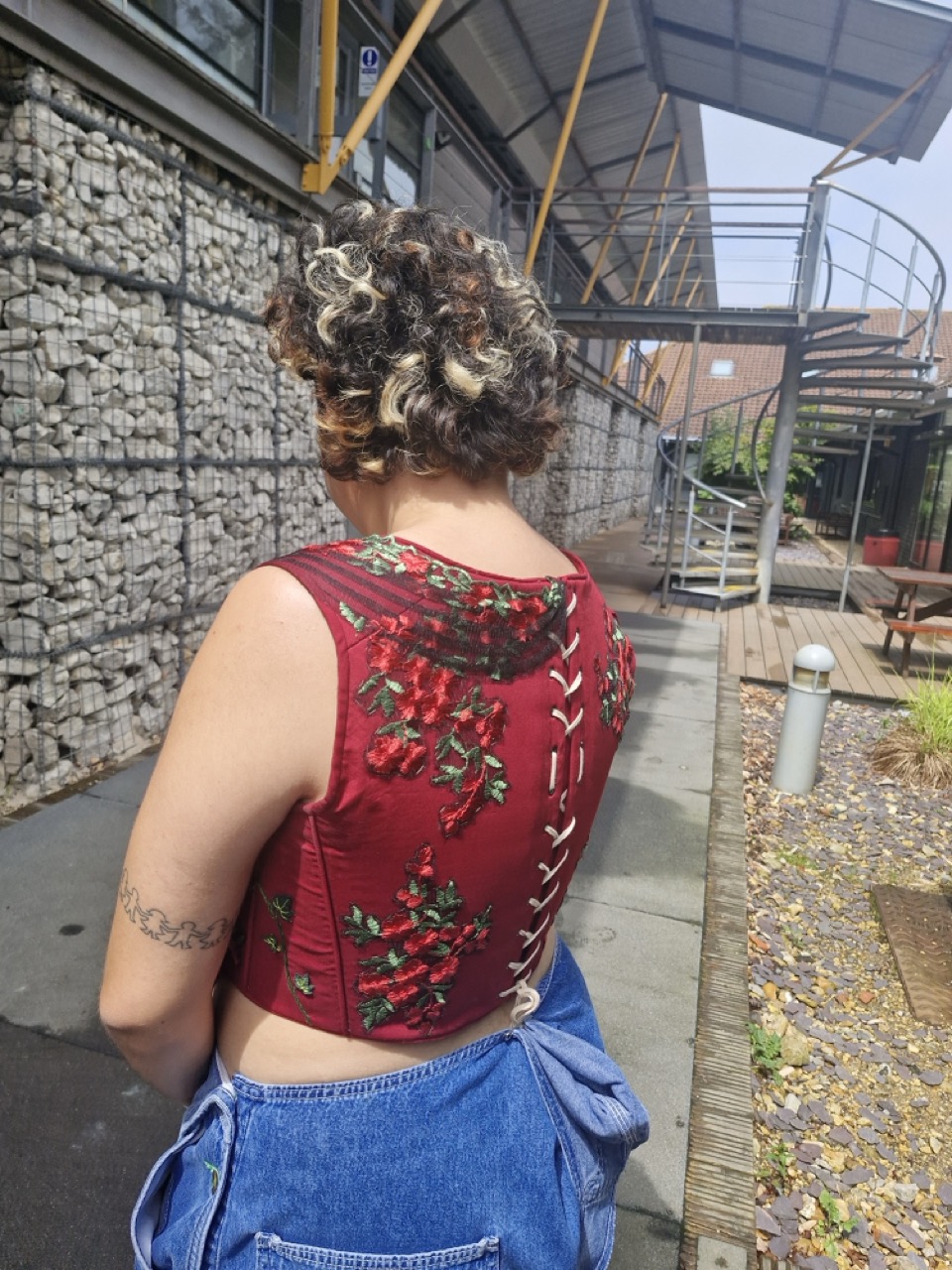 A woman with curly hair wearing a red embroidered crop top with lacing at the back. She is standing outside in a courtyard with a stone wall and a spiral staircase visible in the background.