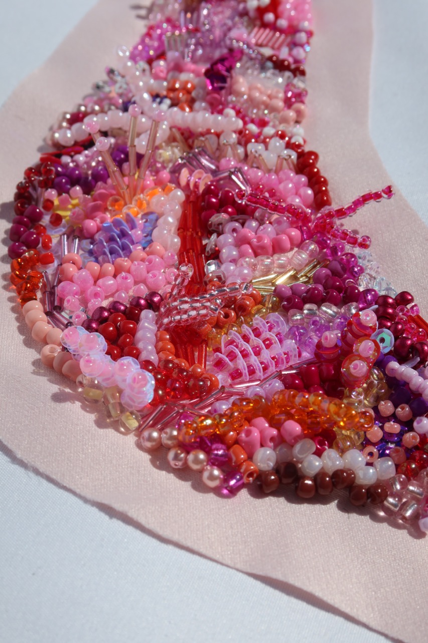 A close-up view of a decorative fabric piece covered in an assortment of multi-coloured beads in shades of pink, red, and orange. The beads are arranged in a flowing pattern on a pale pink backing, showcasing intricate detailing.