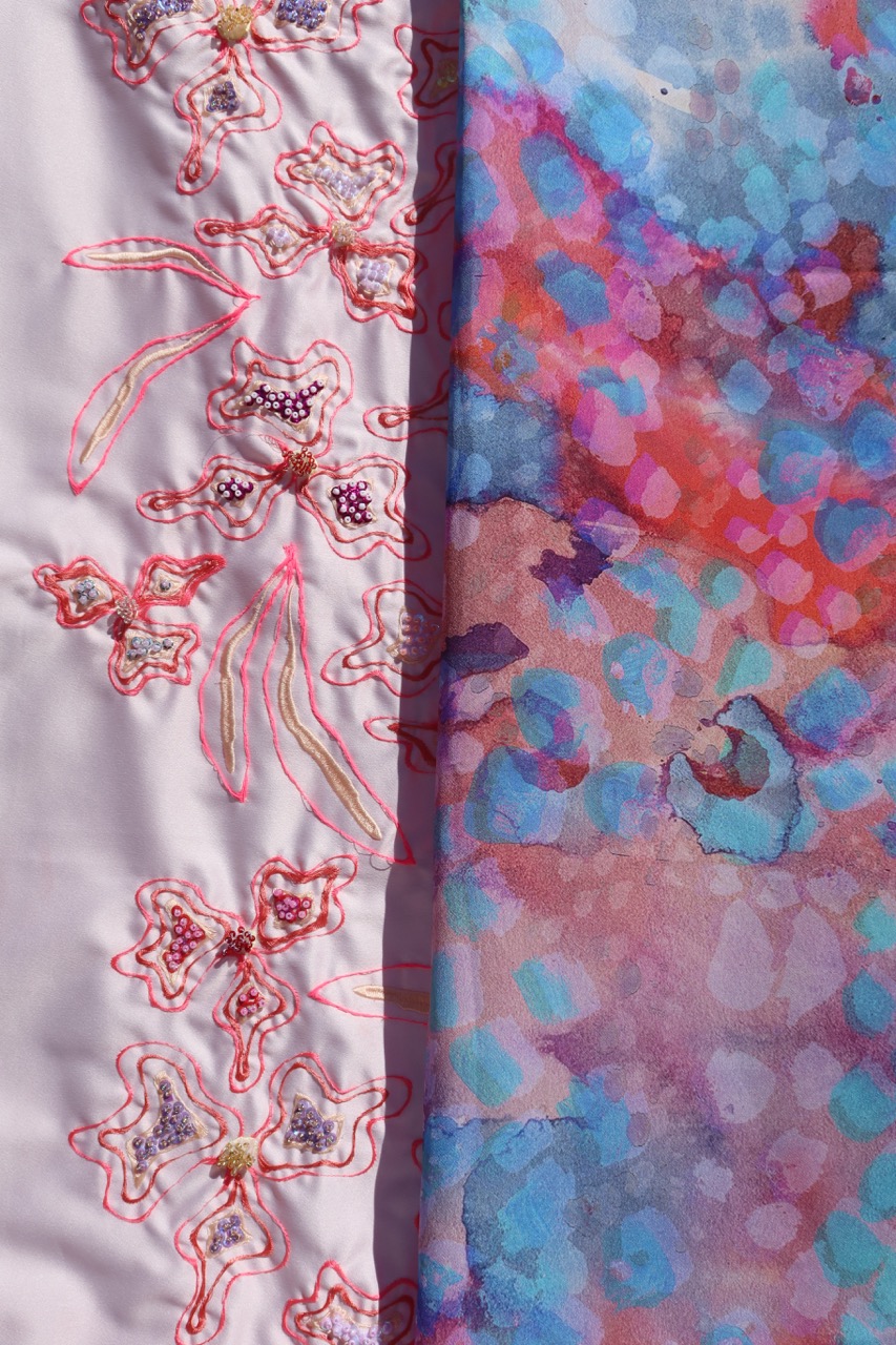 A close-up of fabric showcasing two distinct designs: on the left, embroidered flowers with pink stitching on a light background; on the right, a vibrant, abstract pattern with blue, pink, and purple hues in a dappled arrangement.
