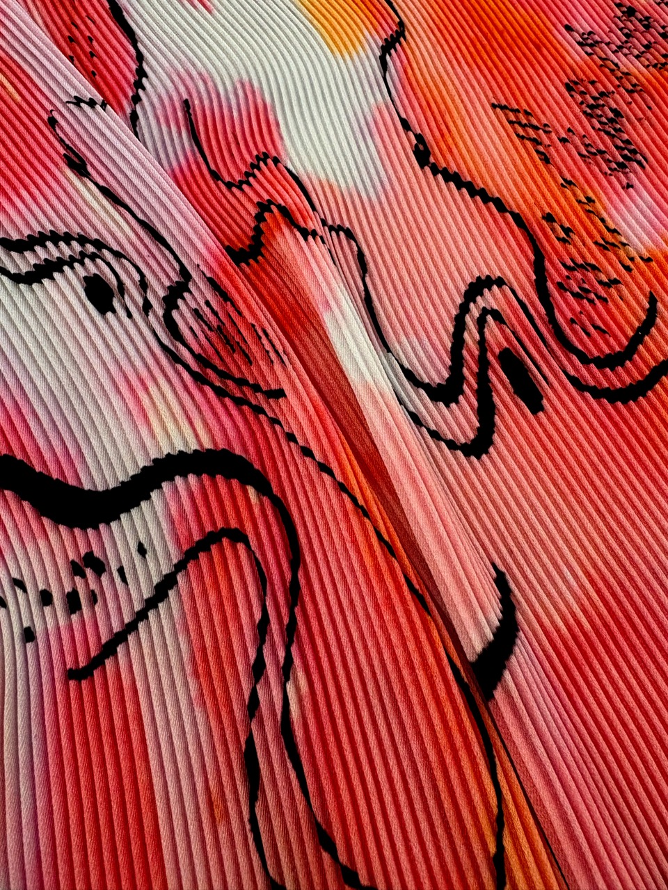 A close-up view of a textured fabric featuring wavy, black lines and abstract shapes, with a gradient of red, pink, and white colours. The fabric has a pleated design that enhances its visual depth.